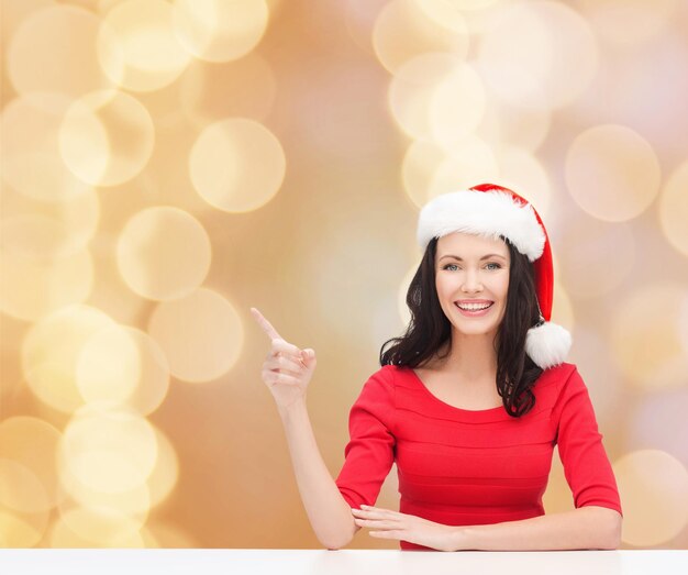 concept de noël, d'hiver, de vacances, de bonheur et de personnes - femme souriante en chapeau de santa helper pointant le doigt sur fond de lumières beiges