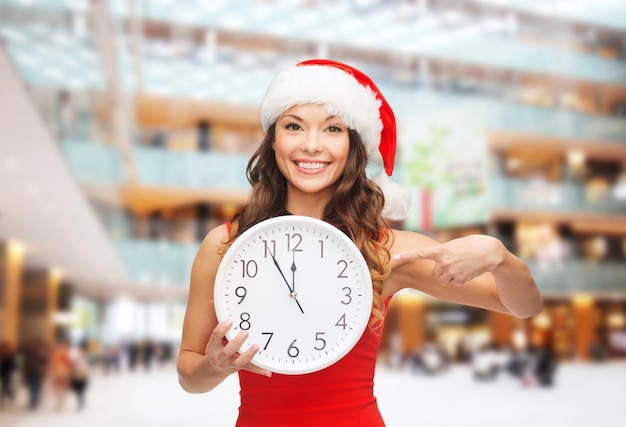 concept de noël, d'hiver, de vacances, de temps et de personnes - femme souriante en bonnet d'assistance et robe rouge avec horloge sur fond de centre commercial