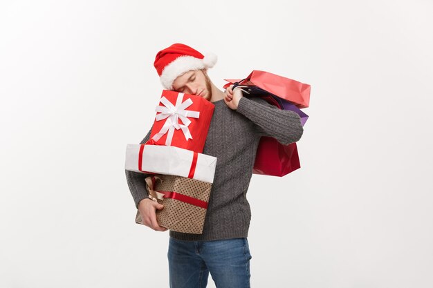 Concept de Noël - jeune bel homme avec barbe tenant des cadeaux lourds avec une expression faciale épuisée sur un mur blanc.