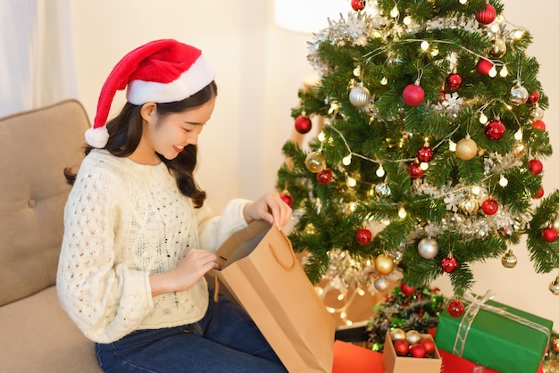 Concept de Noël Jeune femme asiatique en bonnet de Noel souriant et ouvrant le sac cadeau de Noël