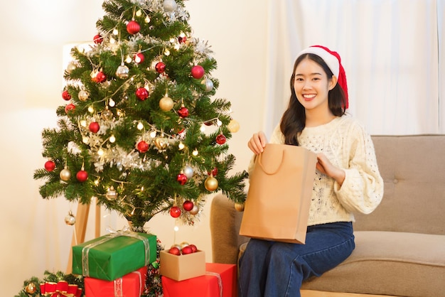 Concept de Noël Jeune femme asiatique en bonnet de Noel souriant et ouvrant le sac cadeau de Noël