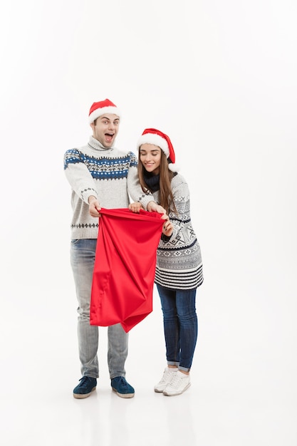Concept de Noël - pleine longueur jeune couple attrayant tenant le sac de santa isolé sur un mur gris blanc.