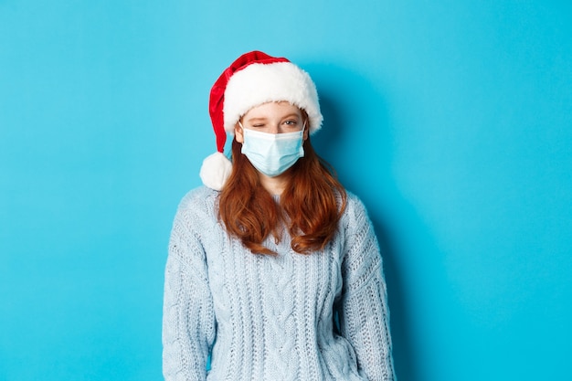 Concept de Noël, de quarantaine et de covid-19. Modèle féminin rousse effronté en masque facial et bonnet de Noel, un clin de œil à la caméra, souhaitant joyeux Noël, debout sur fond bleu.
