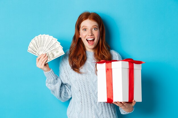 Concept de Noël et shopping. Femme rousse heureuse tenant de l'argent et un grand cadeau de Noël, montrant des dollars et des cadeaux, souriant heureux, debout sur fond bleu.