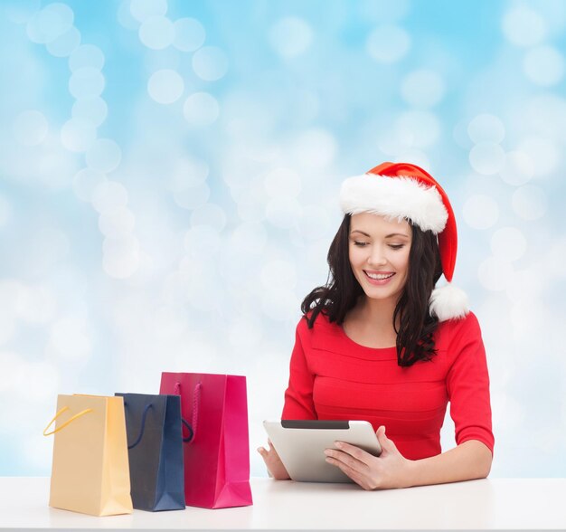 concept de noël, de technologie et de personnes - femme souriante en bonnet d'assistance avec des sacs à provisions et un ordinateur tablette pc sur fond de lumières bleues