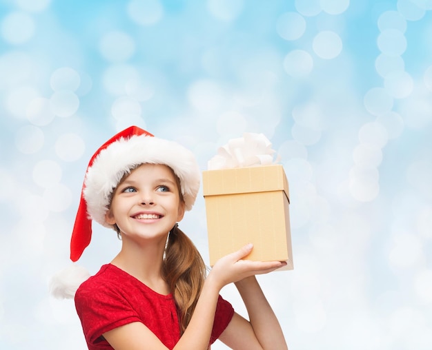 concept de noël, de vacances, d'enfance et de personnes - fille souriante en chapeau de santa helper avec boîte-cadeau sur fond de lumières bleues