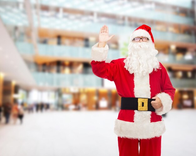 Concept de Noël, de vacances, de gestes et de personnes - homme en costume de père noël agitant la main sur fond de centre commercial