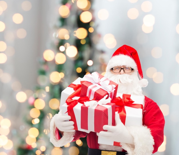 concept de noël, de vacances et de personnes - homme en costume de père noël avec des coffrets cadeaux sur fond de lumières d'arbre