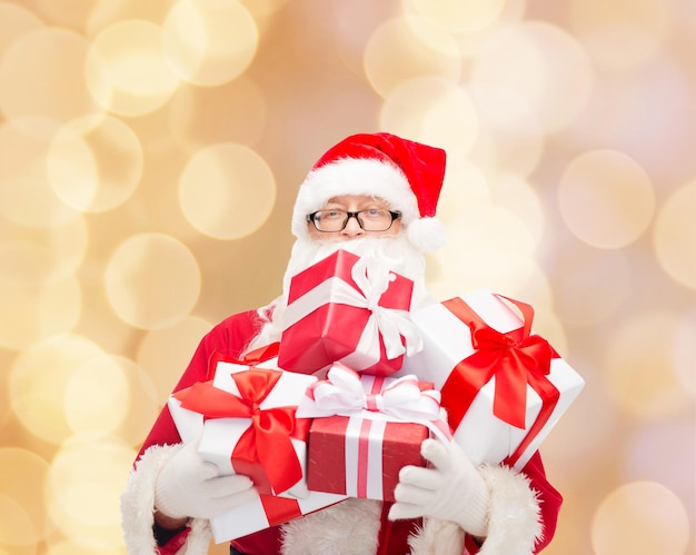 concept de noël, de vacances et de personnes - homme en costume de père noël avec des coffrets cadeaux sur fond de lumières beiges