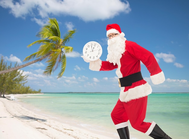 concept de noël, de vacances et de personnes - homme en costume de père noël courant avec horloge montrant douze sur fond de plage tropicale