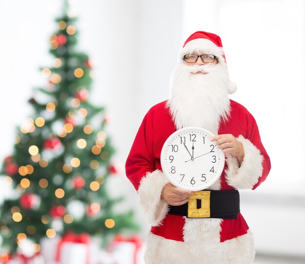 concept de noël, de vacances et de personnes - homme en costume de père noël avec horloge montrant douze doigts pointés sur le salon avec fond d'arbre