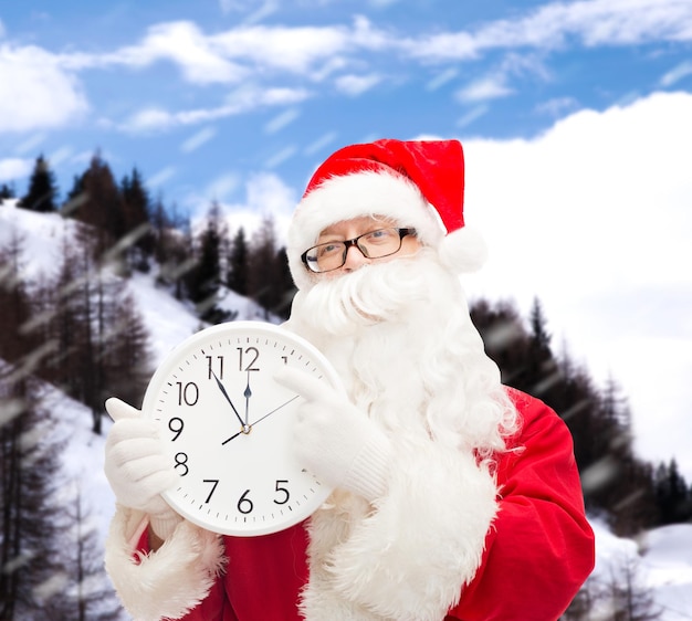 concept de noël, de vacances et de personnes - homme en costume de père noël avec horloge montrant douze sur fond de montagnes enneigées