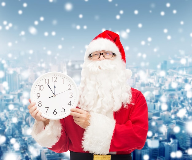 concept de noël, de vacances et de personnes - homme en costume de père noël avec horloge montrant douze sur fond de ville enneigée