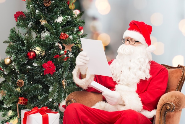 concept de noël, de vacances et de personnes - homme en costume de père noël avec lettre et arbre de noël assis dans un fauteuil sur fond de lumières