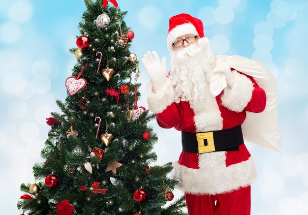 concept de noël, de vacances et de personnes - homme en costume de père noël avec sac et arbre de noël agitant la main sur fond de lumières bleues