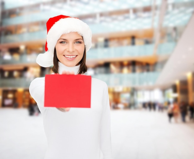 Photo concept de noël, vacances, personnes, publicité et vente - femme heureuse en chapeau de santa helper avec carte rouge vierge sur fond de centre commercial