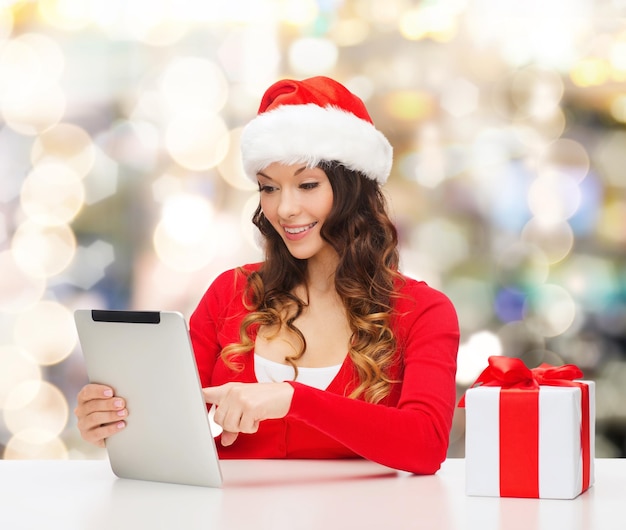 concept de noël, de vacances, de technologie et de personnes - femme souriante en bonnet d'assistance avec boîte-cadeau et ordinateur tablette pc sur fond de lumières