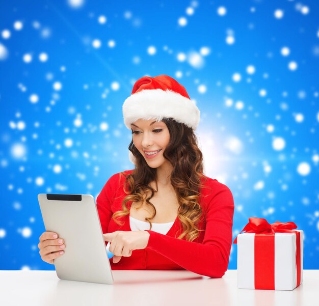 concept de noël, vacances, technologie et personnes - femme souriante en bonnet d'assistance avec boîte-cadeau et ordinateur tablette pc sur fond de neige bleu