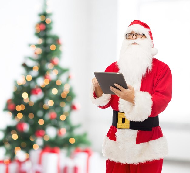 concept de noël, vacances, technologie et personnes - homme en costume de père noël avec tablette pc sur fond de salon et d'arbre