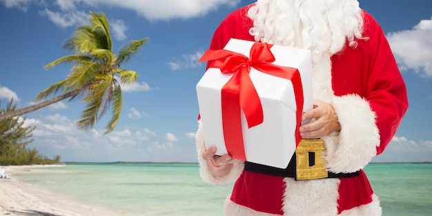 concept de noël, de vacances, de voyage et de personnes - gros plan du père noël avec boîte-cadeau sur fond de plage tropicale