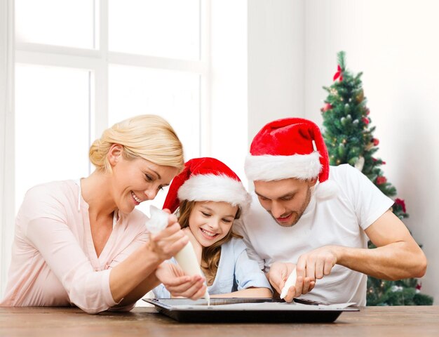 concept de nourriture, de famille, de bonheur et de personnes - famille souriante dans des chapeaux de santa helper avec glaçure et cuisson à la poêle sur fond de salon et d'arbre de noël