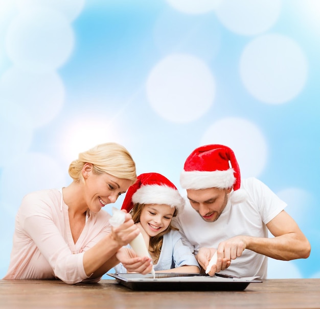 concept de nourriture, de famille, de noël, de bonheur et de personnes - famille souriante dans des chapeaux d'assistant de père Noël décorant des biscuits avec du glaçage