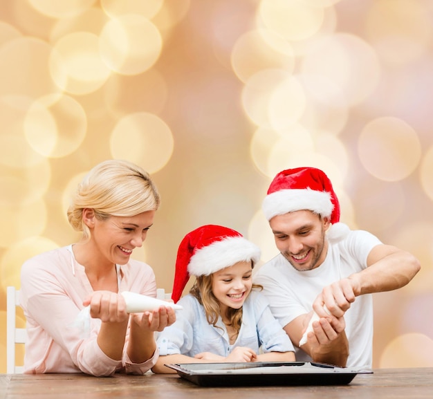 concept de nourriture, de famille, de noël, de bonheur et de personnes - famille souriante dans des chapeaux de santa helper avec glaçure et cuisson à la poêle sur fond de lumières beiges