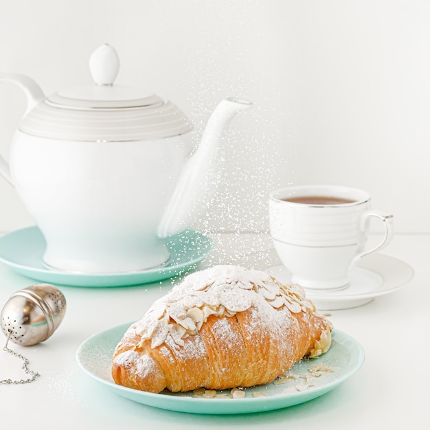 Concept de nourriture volante. Sucre en poudre tombant sur un croissant fraîchement sorti du four sur la table du petit déjeuner blanc avec théière et thé. Image carrée