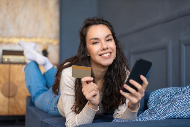 Concept de paiement en ligne femme joyeuse tenant une carte de crédit et un smartphone allongé sur un canapé et souriant