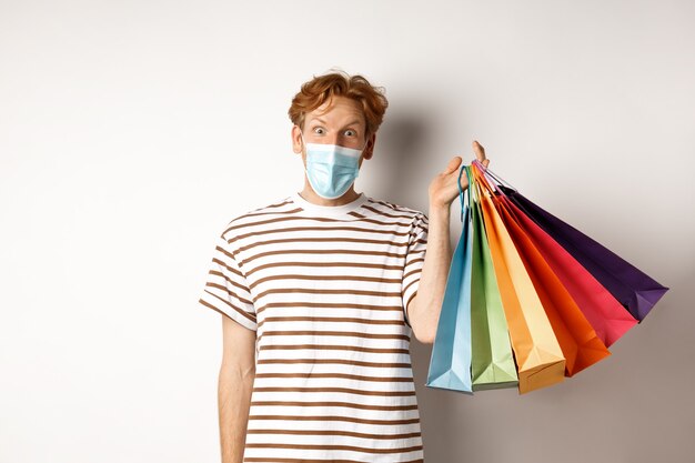 Concept de pandémie et de mode de vie. Beau jeune homme au masque facial étonné de remises spéciales, tenant des sacs à provisions et regardant fond blanc excité.