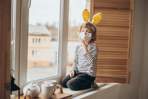 Photo concept de pâques 2021 un enfant en quarantaine à domicile joue près de la fenêtre avec les oreilles d'un lapin sur la tête portant un masque médical contre les virus pendant le coronavirus covid2019 et l'épidémie de grippe