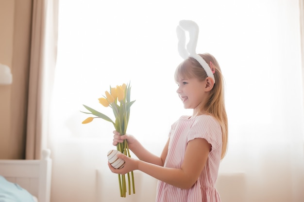 Concept de Pâques. Petite fille souriante en oreilles de lapin blanc moelleux et robe rose tenant des fleurs de tulipes jaunes et oeuf coloré. Intérieur de la chambre.