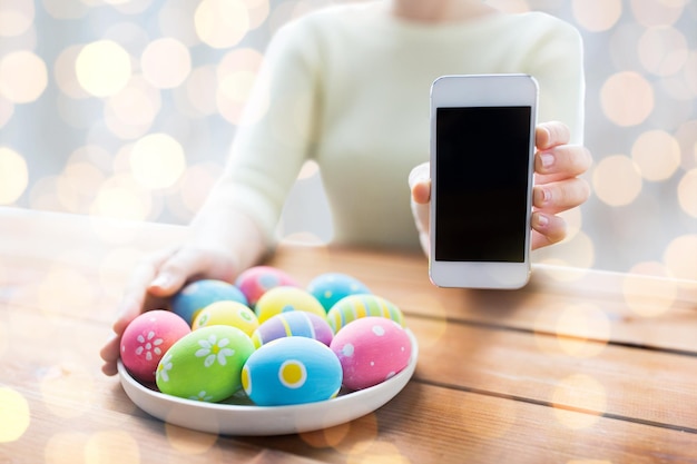 concept de pâques, de vacances, de technologie et de personnes - gros plan sur des mains de femme avec des oeufs de pâques colorés sur une assiette et un smartphone