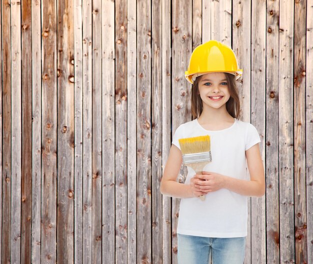 concept de peinture, de construction, d'enfance et de personnes - petite fille souriante dans un casque de protection avec un pinceau sur fond de clôture en bois