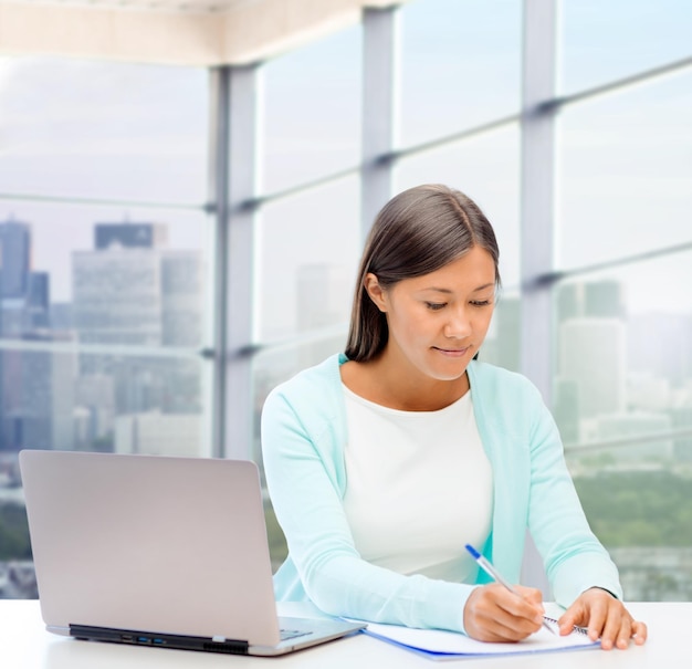concept de personnes, d'affaires et de technologie - femme souriante avec un ordinateur portable écrivant sur un ordinateur portable sur fond de fenêtre de bureau