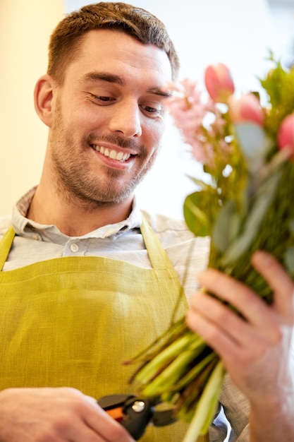 concept de personnes, d'affaires, de vente et de fleuristerie - homme fleuriste souriant et heureux faisant des bouquets et recadrant des tiges par des ciseaux au magasin de fleurs