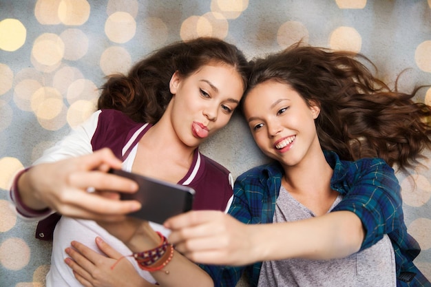 concept de personnes, d'amis, d'adolescents et d'amitié - jolies adolescentes souriantes et heureuses allongées sur le sol et prenant un selfie avec un smartphone sur fond de lumières de vacances