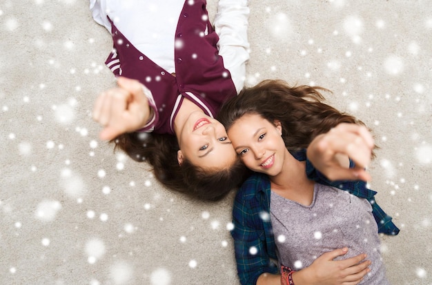 concept de personnes, d'amis, d'hiver, de noël et d'amitié - jolies adolescentes souriantes et heureuses allongées sur le sol et pointant le doigt vers vous sur la neige