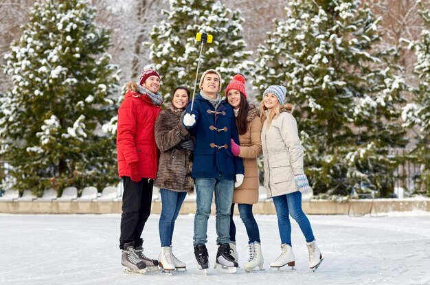 concept de personnes, d'amitié, de technologie et de loisirs - amis heureux prenant des photos avec un bâton de selfie de smartphone sur une patinoire à l'extérieur
