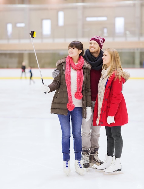 concept de personnes, d'amitié, de technologie et de loisirs - amis heureux prenant des photos avec un bâton de selfie de smartphone sur une patinoire