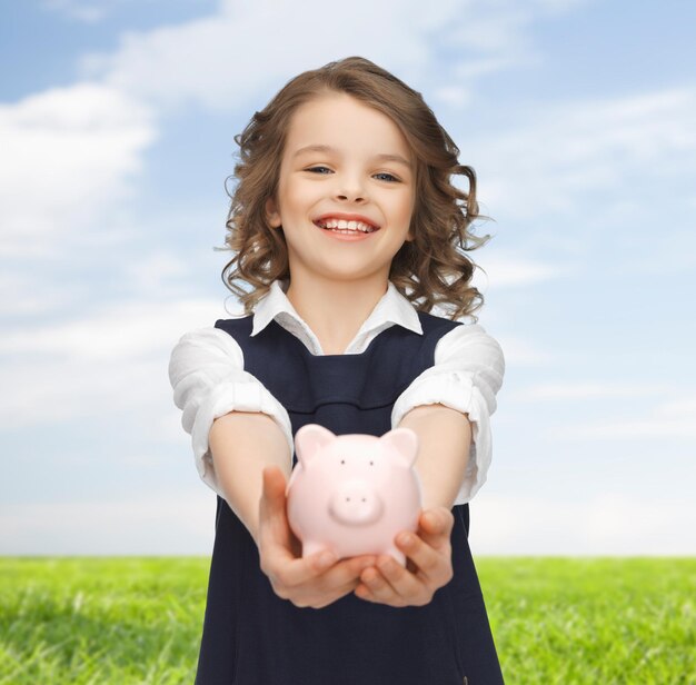 concept de personnes, d'argent, de finances et d'épargne - fille heureuse tenant une tirelire sur les paumes sur fond de ciel bleu et d'herbe