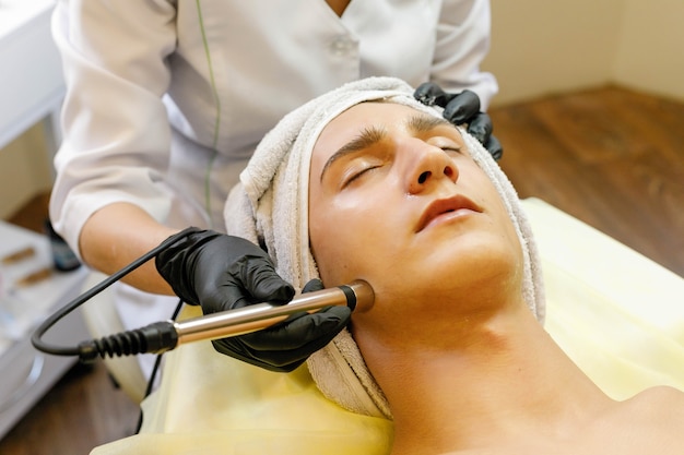 Concept de personnes, de beauté, de spa, de cosmétologie et de technologie - Le cosmétologue effectue la thérapie par micro-courant de la peau du visage d'un beau jeune homme dans un salon de beauté