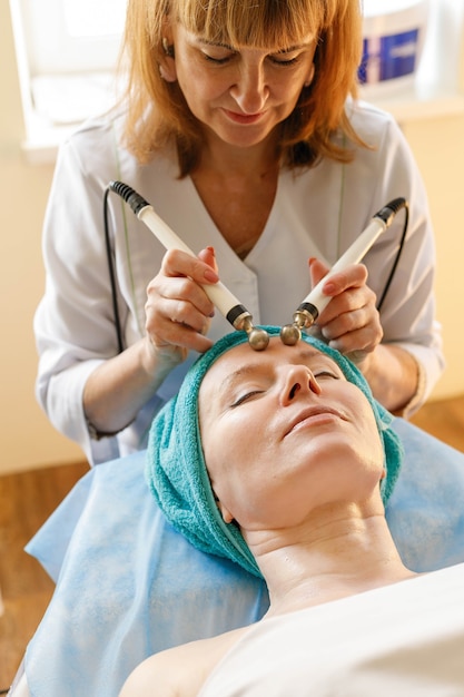 Concept de personnes, de beauté, de spa, de cosmétologie et de technologie - Le cosmétologue effectue la thérapie par micro-courant de la peau du visage d'une belle jeune femme dans un salon de beauté