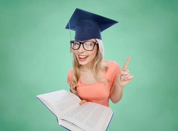 concept de personnes, d'éducation, de connaissances et de remise des diplômes - jeune étudiante souriante dans un mortier et des lunettes avec un livre d'encyclopédie pointant le doigt vers le haut sur l'arrière-plan du tableau de craie de l'école verte