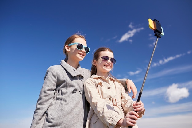 concept de personnes, d'enfants, d'amis et d'amis - filles heureuses prenant des photos avec un smartphone sur un bâton de selfie à l'extérieur