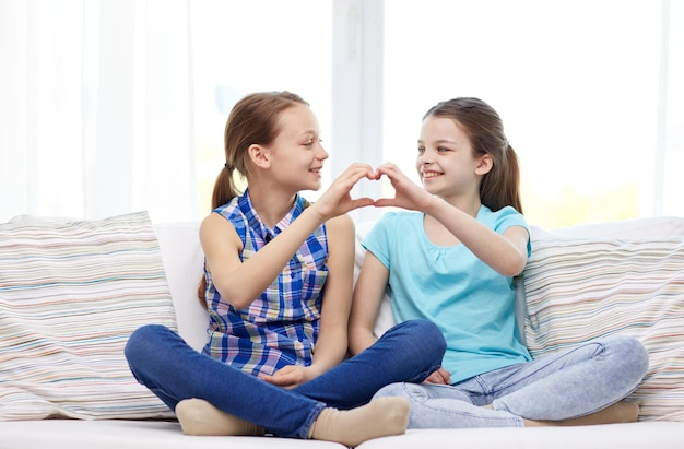 concept de personnes, d'enfants, d'amis et d'amitié - petites filles heureuses assises sur un canapé et montrant un signe de main en forme de coeur à la maison