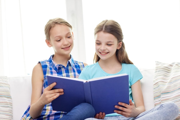 concept de personnes, d'enfants, d'amis, de littérature et d'amitié - deux filles heureuses assises sur un canapé et lisant un livre à la maison