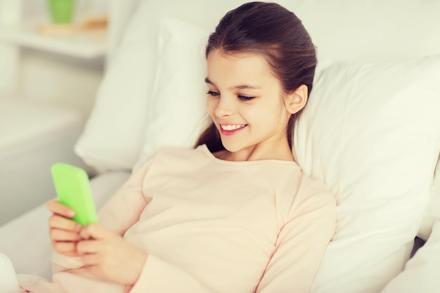 concept de personnes, d'enfants, de repos et de technologie - heureuse fille souriante allongée éveillée avec un smartphone au lit à la maison