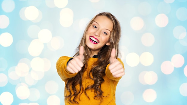 concept de personnes, de gestes, de style et de mode - heureuse jeune femme ou adolescente en vêtements décontractés montrant les pouces vers le haut sur fond de lumières bleues de vacances