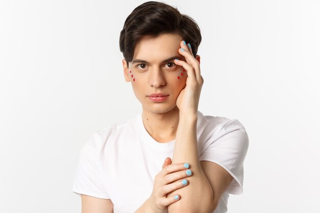 Concept de personnes, de lgbtq et de beauté. Gros plan du bel homme queer touchant le visage avec les doigts avec du vernis à ongles bleu, debout sur blanc.